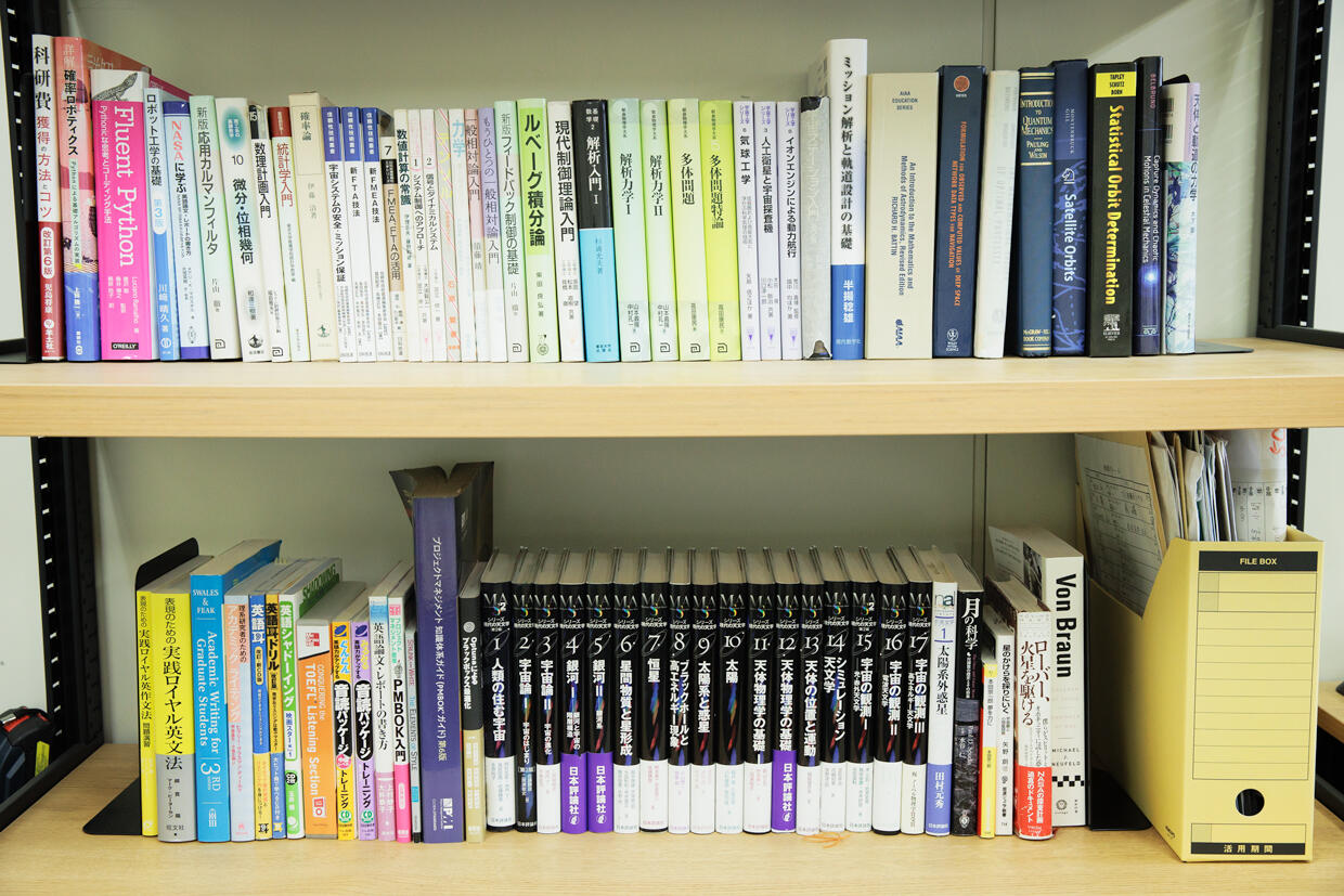Ozaki's bookshelf in the laboratory. The upper shelf contains books on physics and mathematics essential for trajectory design, such as analytical mechanics, N-body problems, and probability theory, while the lower shelf contains a variety of books to gain broad knowledge for mission design, such as the “Modern Astronomy Series.”
